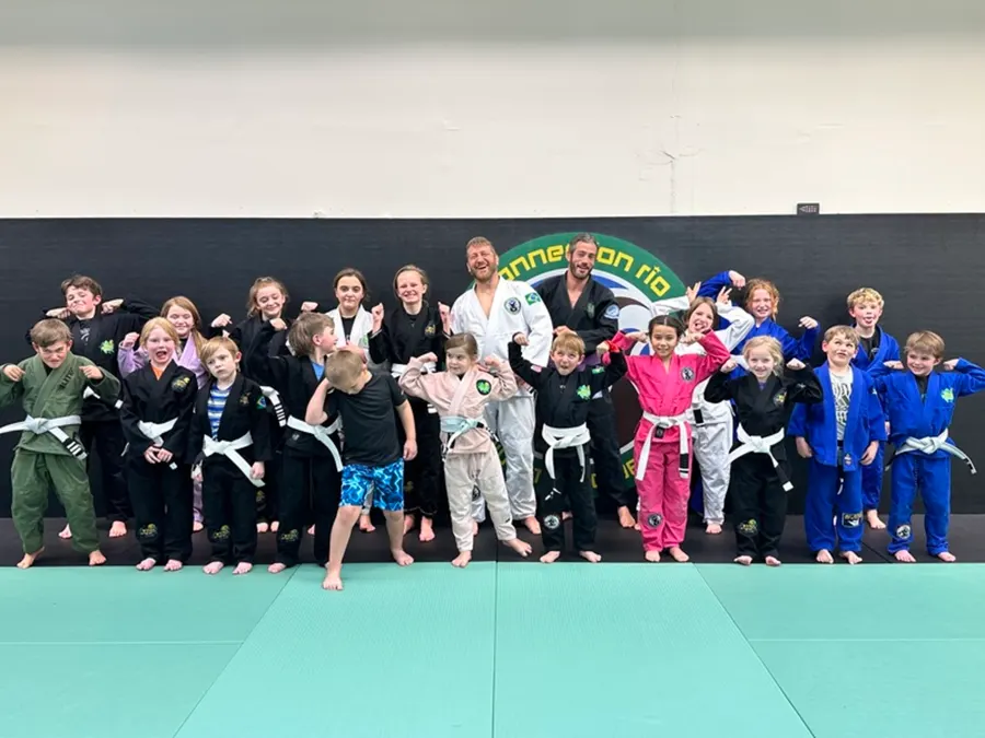 A big group of kids in gis and rash guards posing on green mats in front of the Connection Rio logo