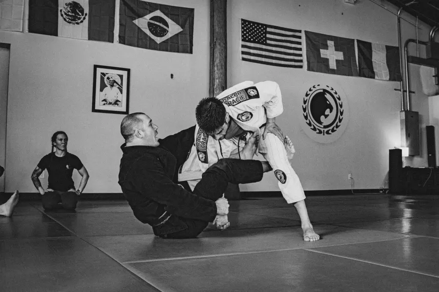 Black and white photo of a student in a white gi passing a partner’s guard under Brazilian and U.S. flags at Ralph Gracie Bend