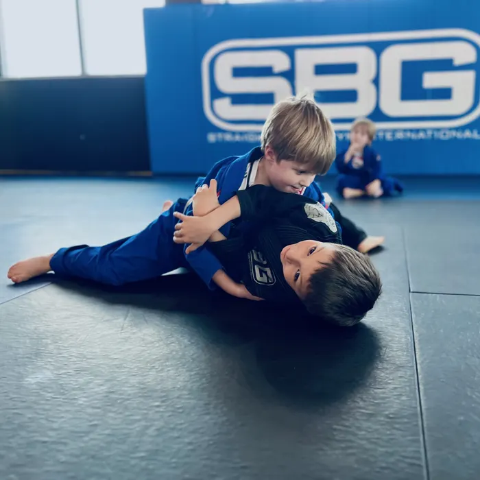 Two young kids in blue gis grappling on the mat in front of an SBG banner