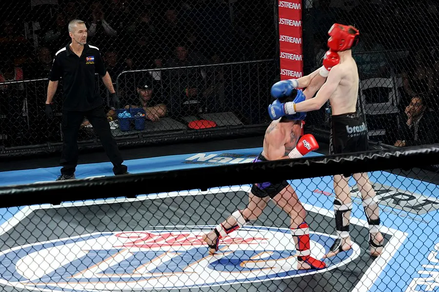 A referee watches two amateur MMA fighters in headgear exchange punches inside a cage