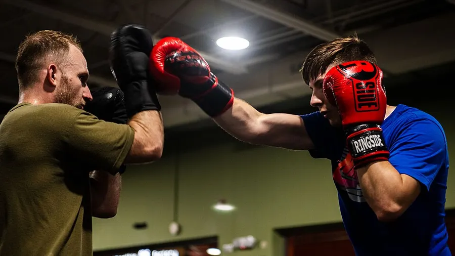 Two boxers in gloves work a punch-and-block drill in a gym with heavy bags behind them