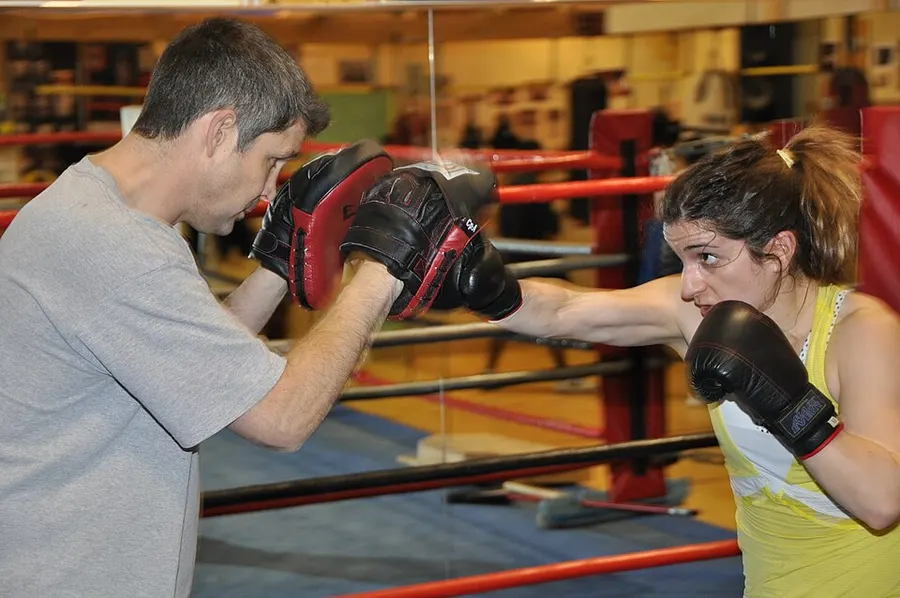 A female fighter in boxing gloves punches focus mitts held by her coach inside a boxing ring