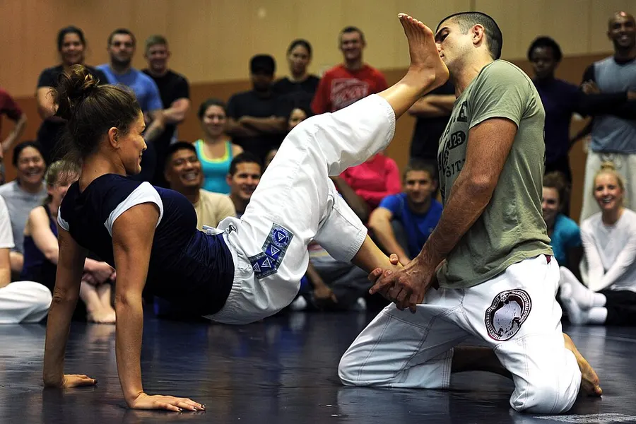 A female instructor on the mat demonstrates a self-defense kick on a kneeling partner while a class watches