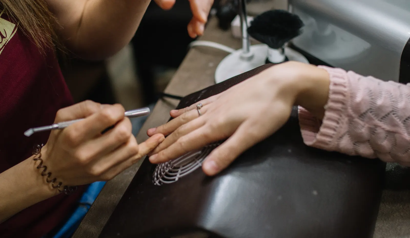 A nail technician works on a client's cuticles at a manicure table under a desk lamp