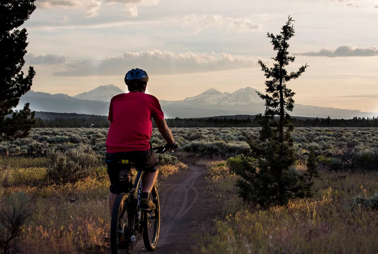 A mountain biker in a red jersey rides a sagebrush singletrack at Cline Buttes with the snowy Three Sisters on the horizon