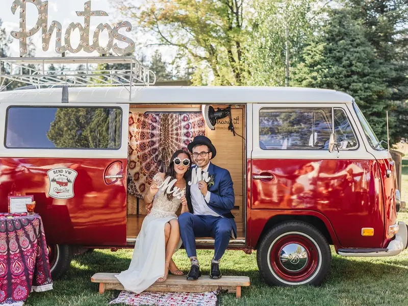 A newly married couple sitting in the open door of a red-and-white vintage VW bus photo booth on a lawn