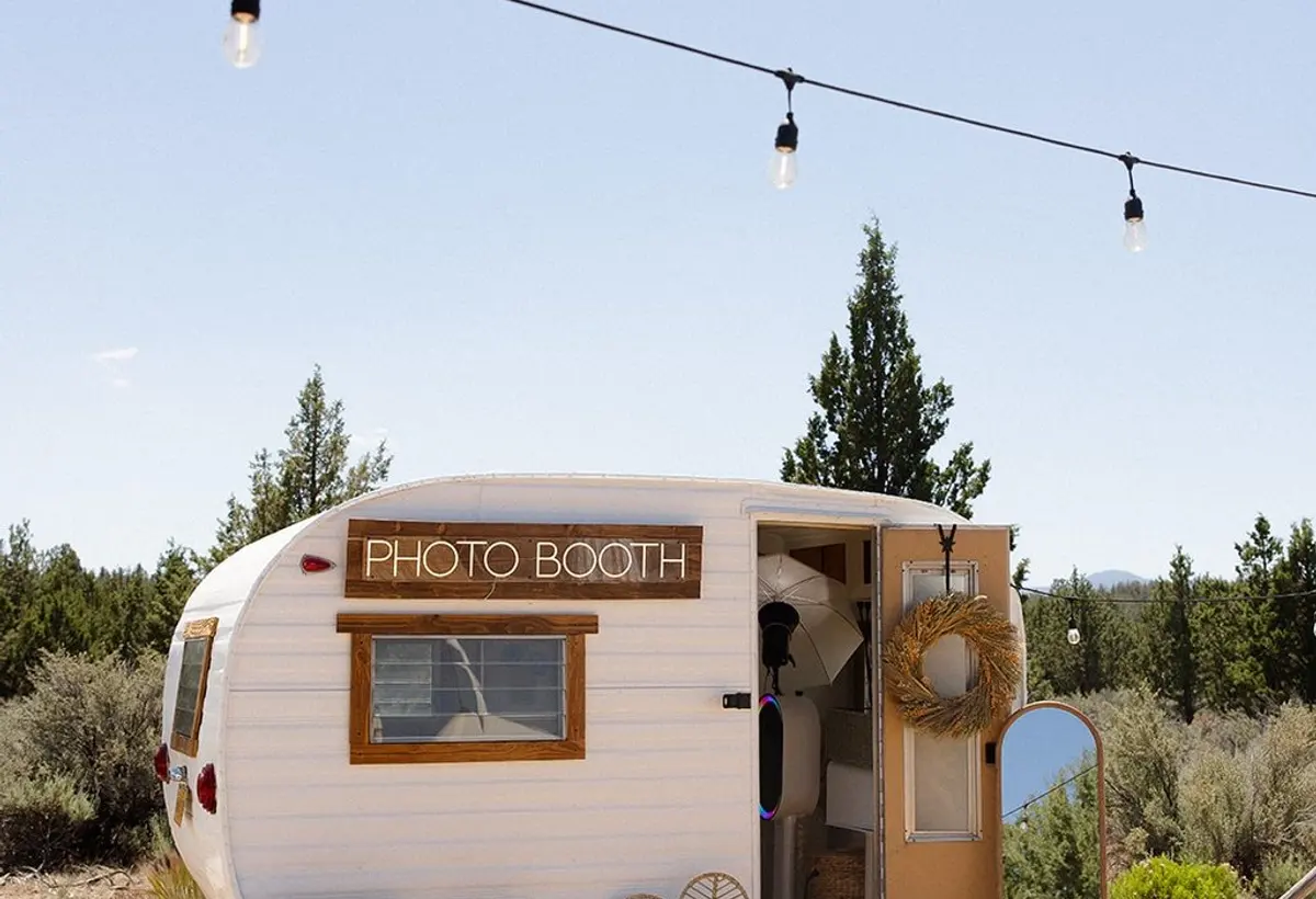 A restored white 1962 camper trailer with a Photo Booth sign, string lights and lounge chairs on a high-desert lot with junipers