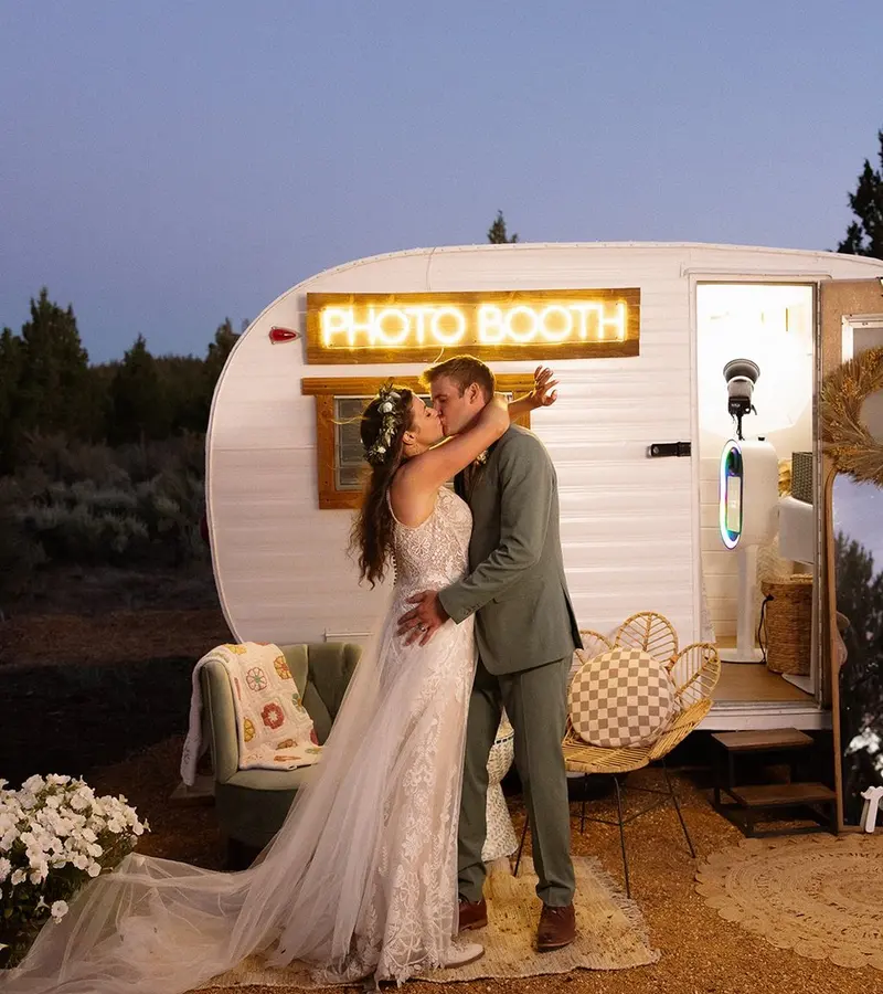 A bride and groom kissing in front of a white vintage camper with a glowing Photo Booth sign at dusk among junipers