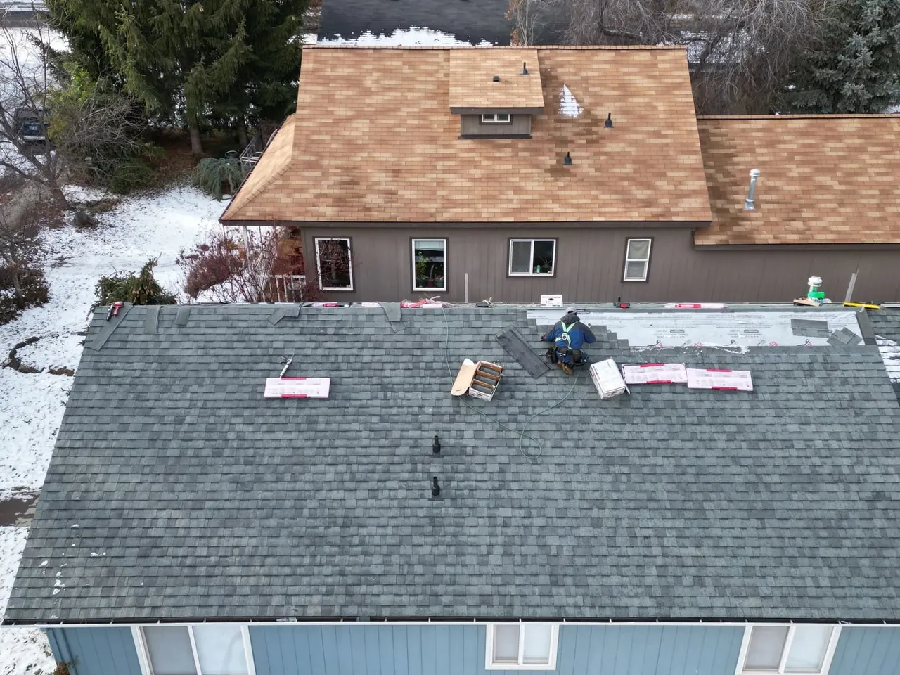 Aerial view of a roofer working on a dark shingle roof in Bend with snow on the ground and a brown shingle roof behind