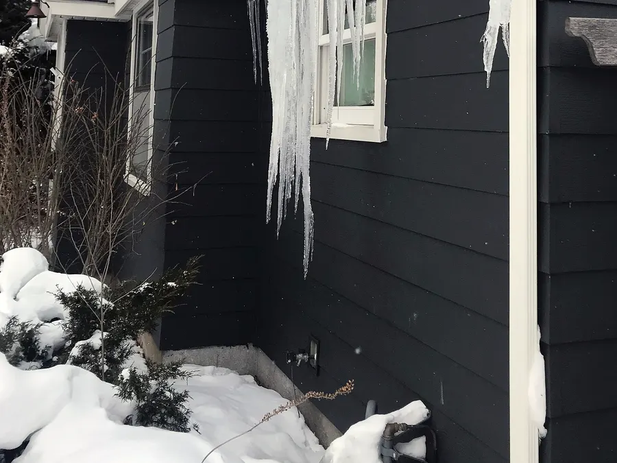 A thick curtain of icicles hanging from snowy eaves down past a window on a dark-sided house
