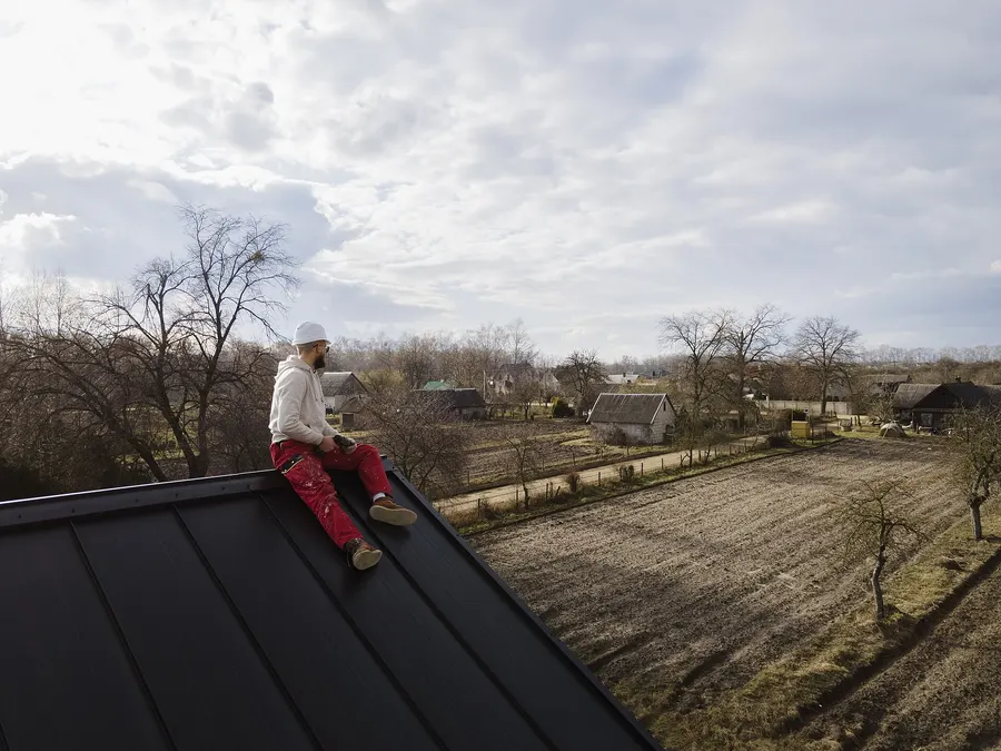 A roofer in a white hard hat sitting at the ridge of a dark standing seam metal roof above farm fields