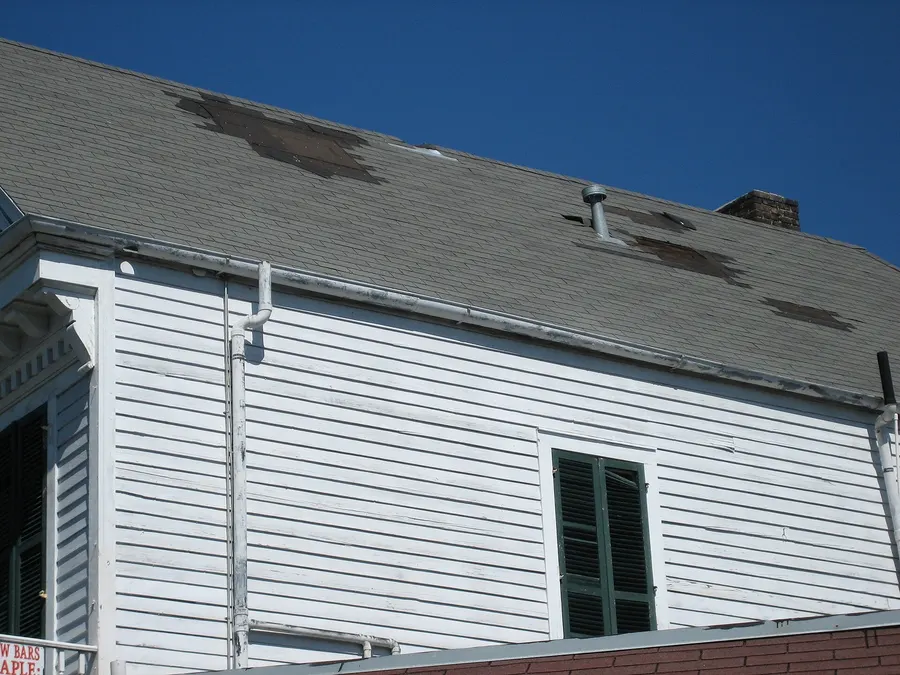 A gray shingle roof on a white house with dark patches where wind tore shingles off