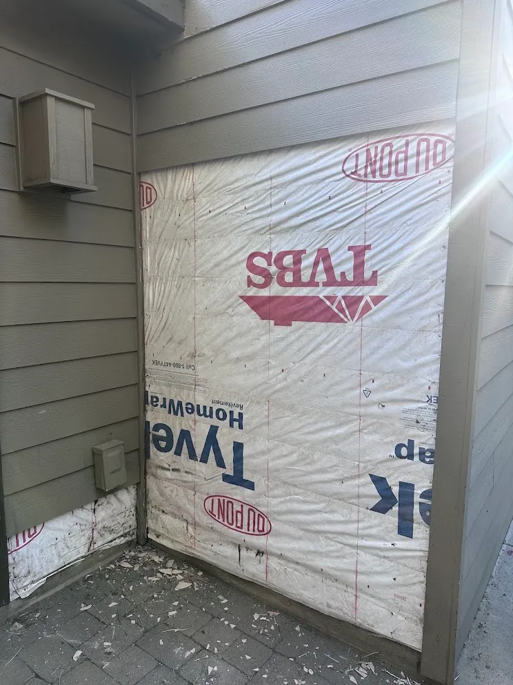 Exterior wall with the old lap siding removed, showing the Tyvek house wrap underneath and broken siding pieces on the pavers