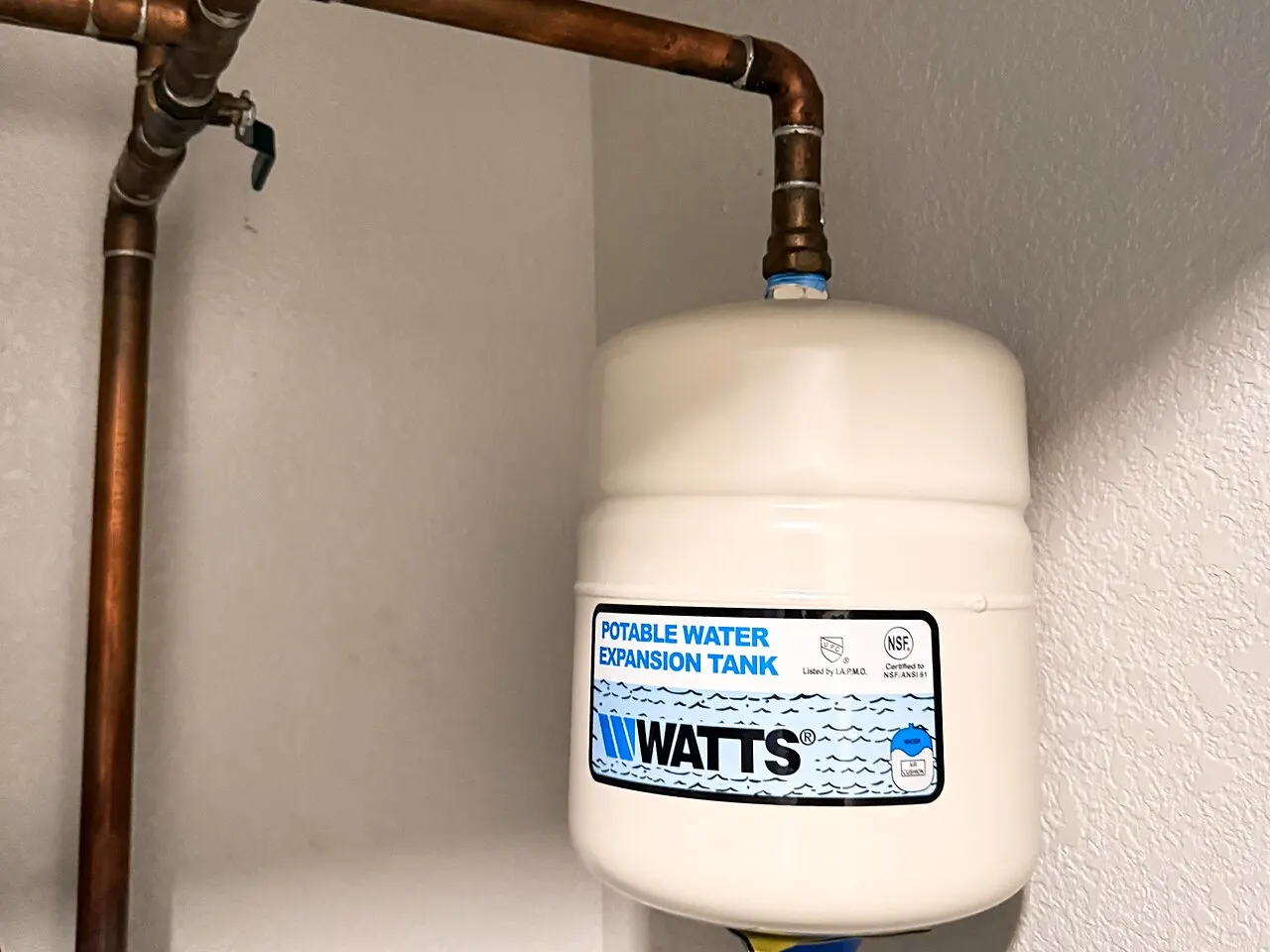 A white Watts potable water expansion tank hanging from copper pipes above a water heater