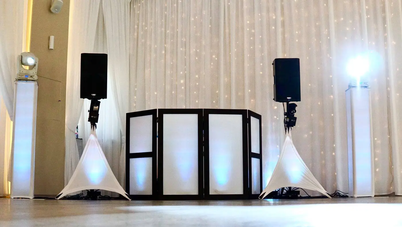 A white DJ booth with speakers on stands and blue uplighting against a curtain of string lights at a wedding reception