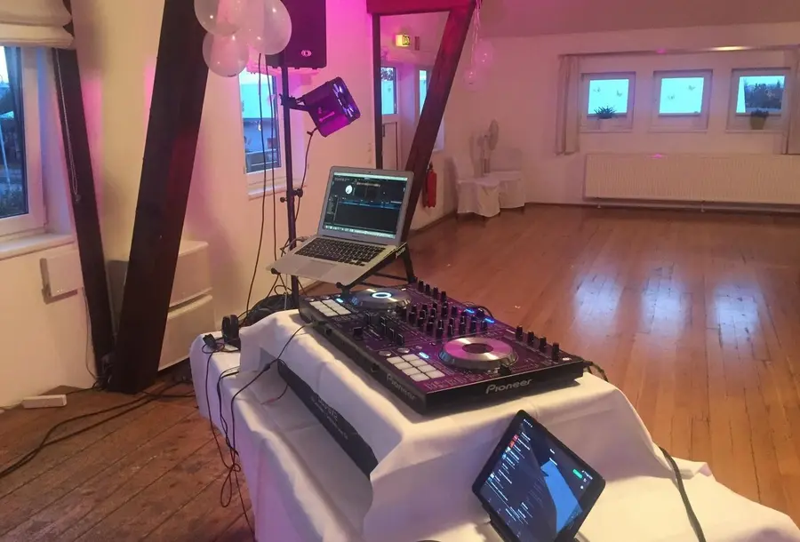 A DJ controller and laptop on a white-draped table in a wedding reception room