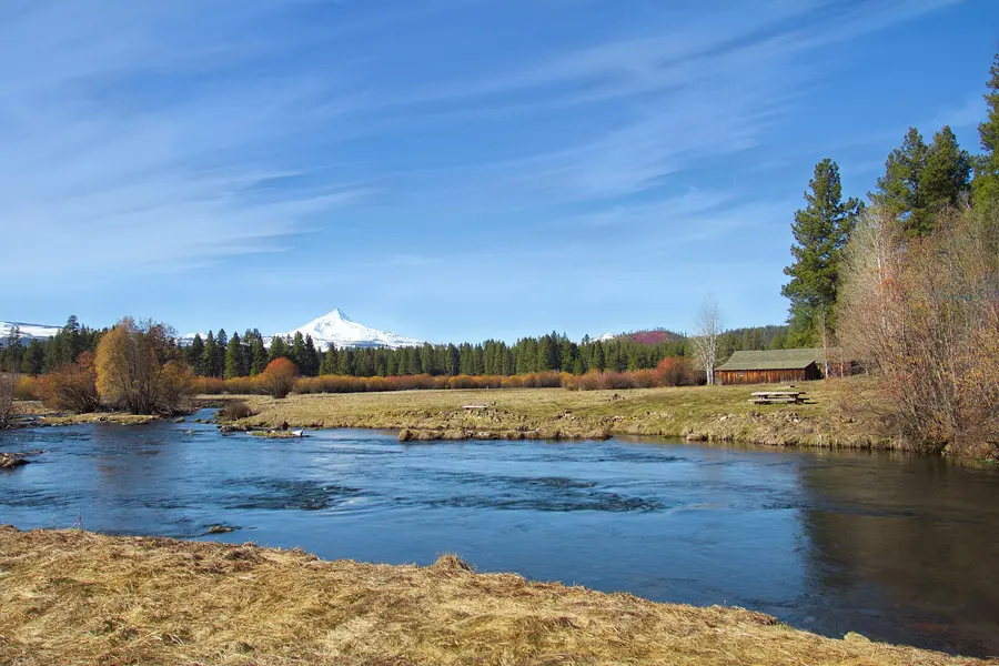 The clear Metolius River flowing past a meadow and old barn, with snowy Mt. Jefferson beyond the pines