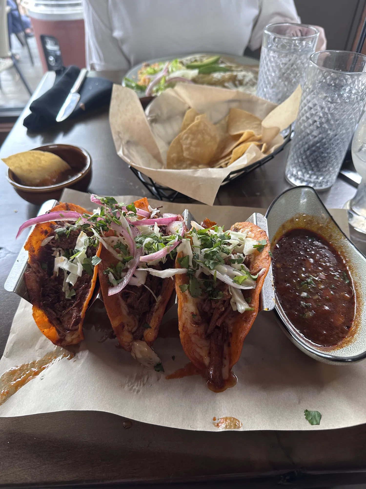 Three birria tacos with onion and cilantro next to a cup of consomé