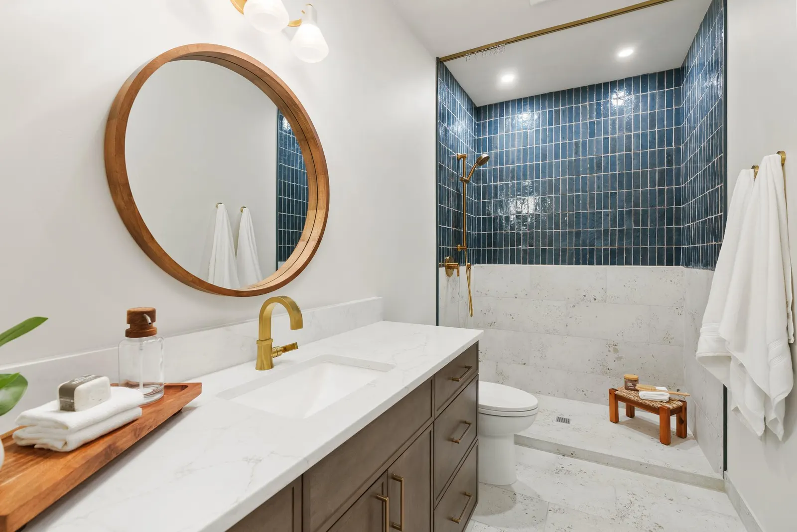 Guest bath with a round wood mirror, white quartz vanity and blue tile shower by Surface Kitchen & Bath in Bend