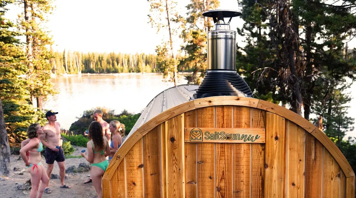 A cedar mobile barrel sauna with a stovepipe beside a mountain lake