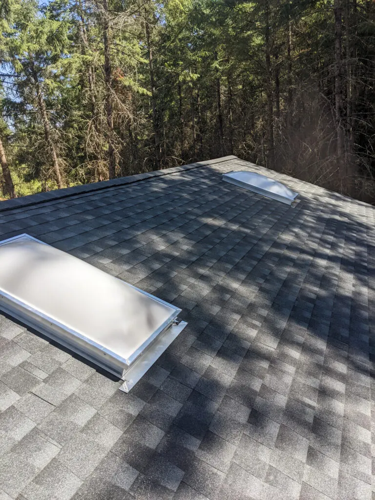 A domed skylight on a gray shingle roof surrounded by pine trees, installed by RCO Roofing