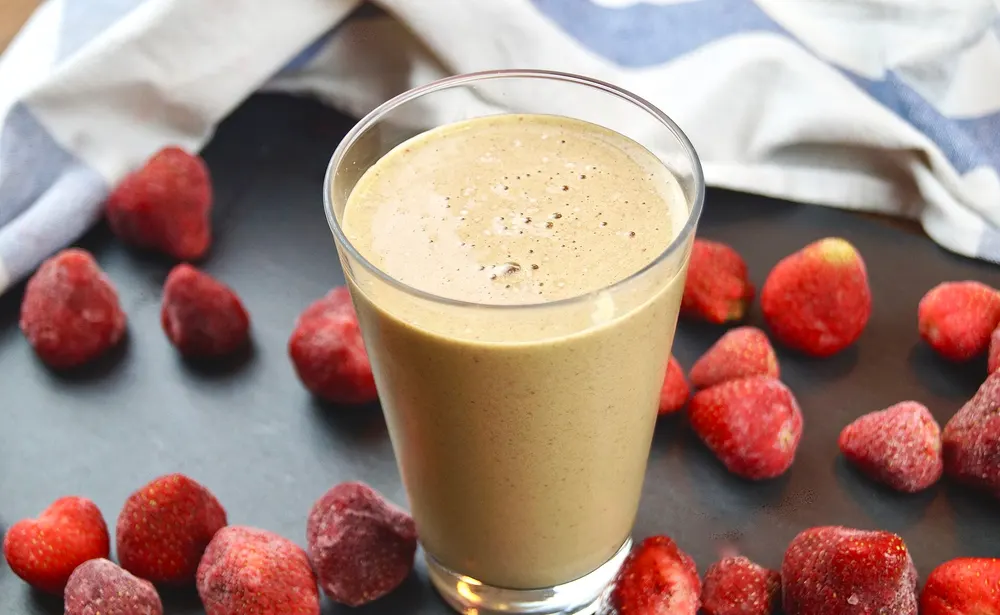 A glass of strawberry smoothie surrounded by frozen strawberries