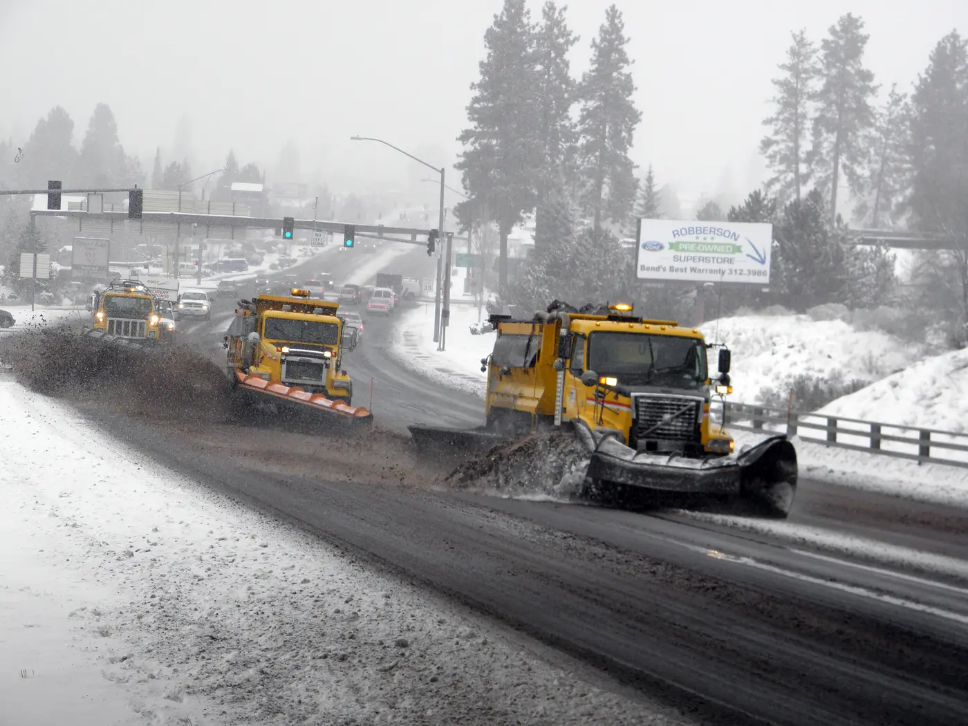 Three yellow ODOT snowplows clearing U.S. 20 through snowy pines in Bend