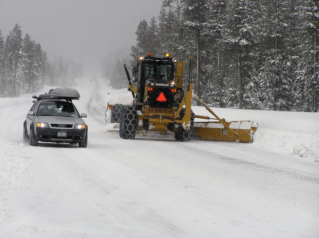 A road grader with chains plowing snowy Century Drive past a car with a roof box, between snow-covered pines
