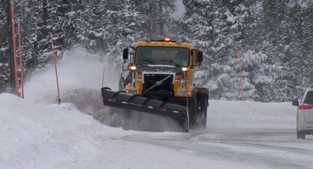 A snowplow throwing a wave of snow past orange marker poles in a snowy forest
