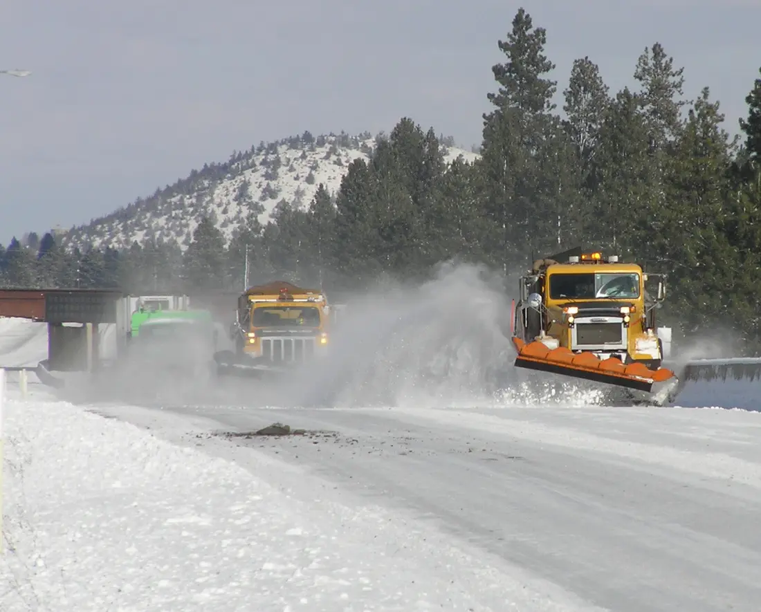 ODOT snowplows throwing snow off U.S. 97 in Bend, with a pine-covered butte behind