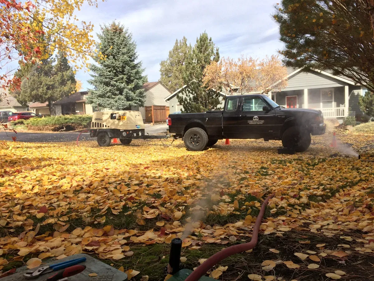 An irrigation service truck blowing out sprinklers on a big yard covered in fall leaves