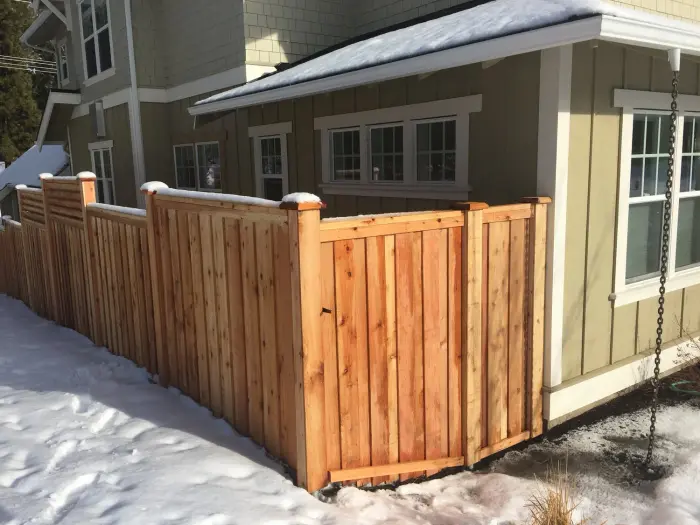 A new cedar good-neighbor fence with stepped top caps beside a gray house, with snow on the ground
