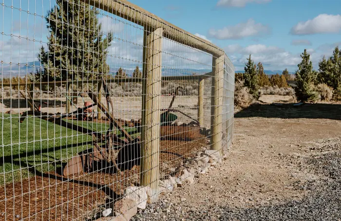 A tall wood-post fence with welded wire panels around a garden on open high-desert land with junipers