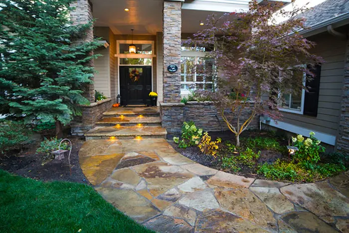 A flagstone walk and steps leading to a lit front door with stone columns and planting beds