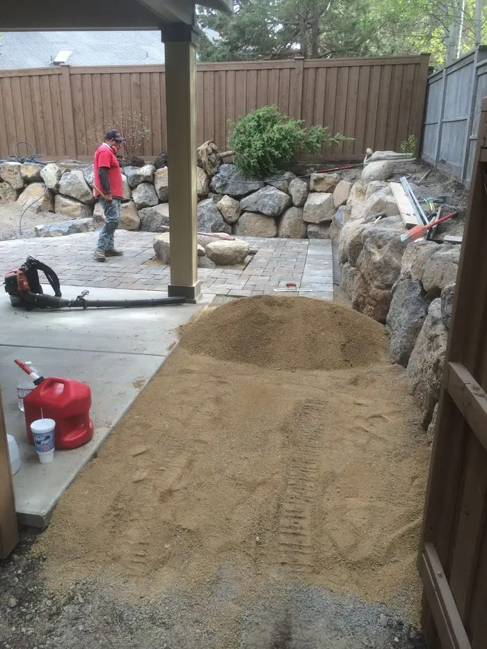 A crew screeding bedding material and compacting base for a paver patio beside a basalt boulder wall