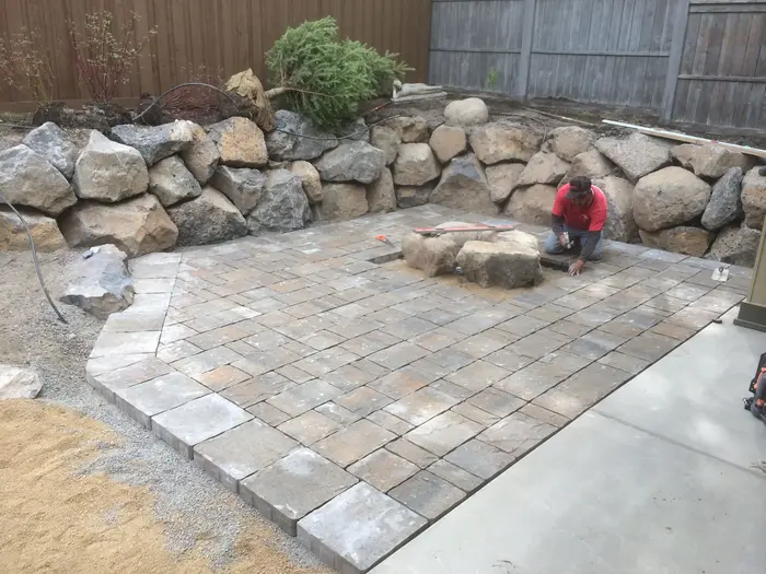 A worker setting gray concrete pavers around a boulder fire pit ring inside a basalt boulder wall