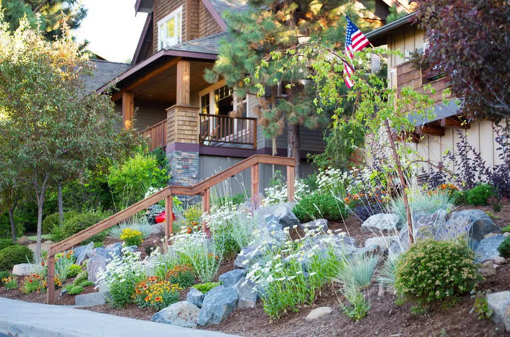 A front-yard xeriscape of boulders, ornamental grasses and flowering perennials climbing to a craftsman house under pines on Awbrey Butte
